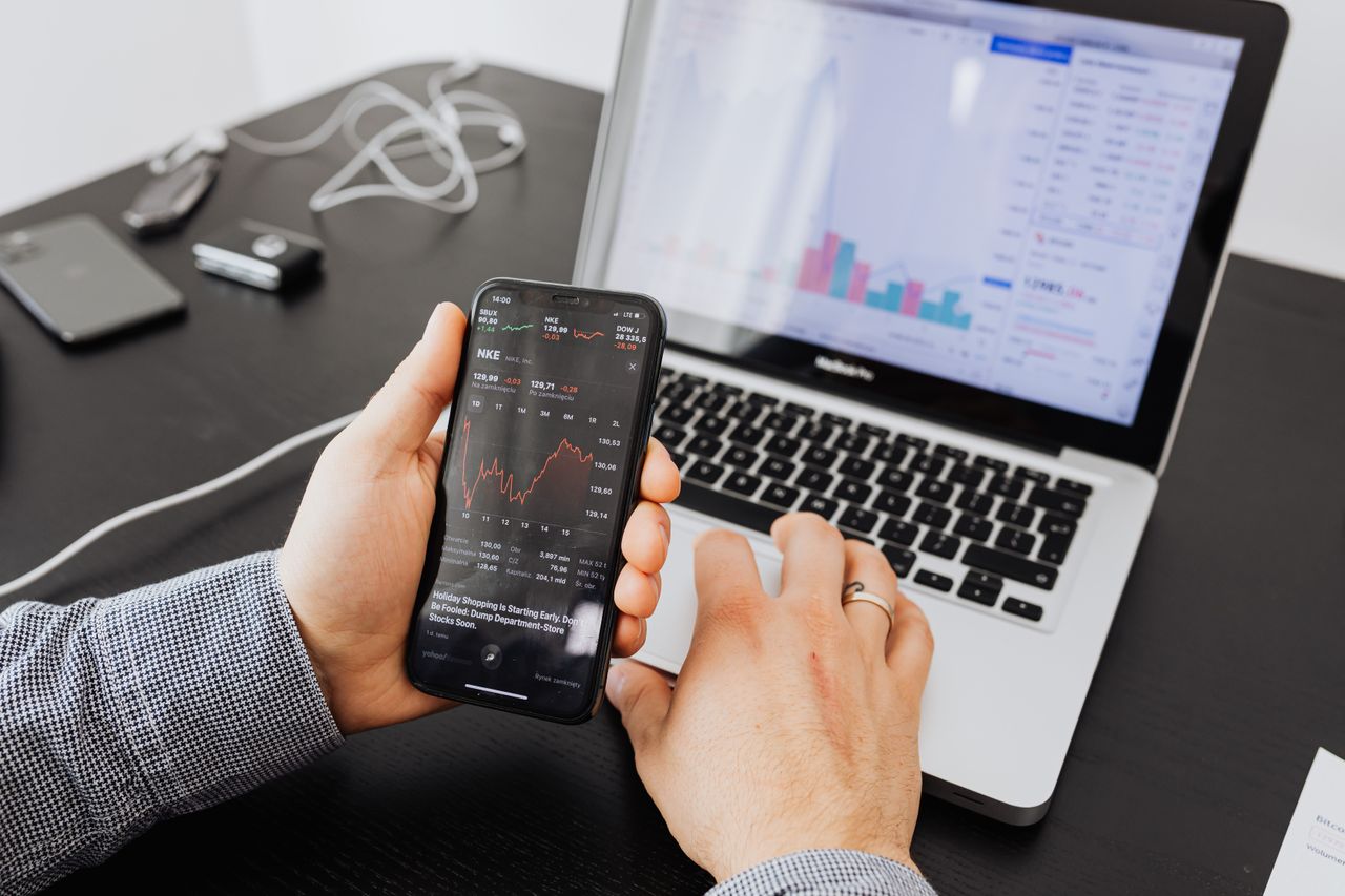 Personne analysant des données boursières avec un smartphone et un ordinateur portable, à l’intérieur.